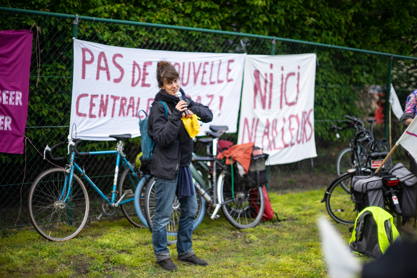 Activiste met microfoon staat voor een omheining waarop spandoeken hangen "pas de nouvelle centrale de gaz!" en waartegen fietsen leunen
